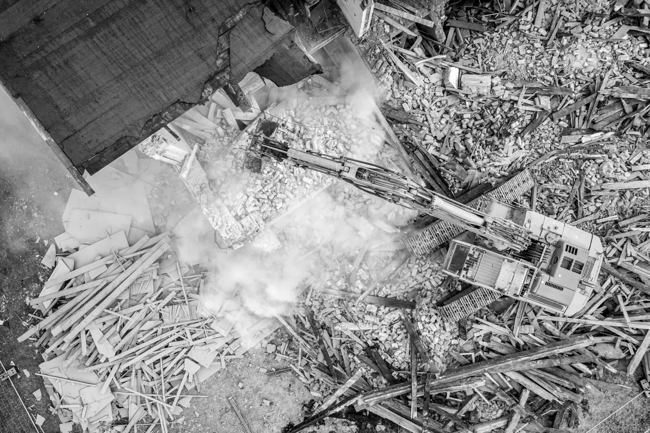 Abbrucharbeiten aus der Luft: Bagger mit Abbruchgreifer räumt ein niedergelegtes Gebäude, Staub steht über der Baustelle.